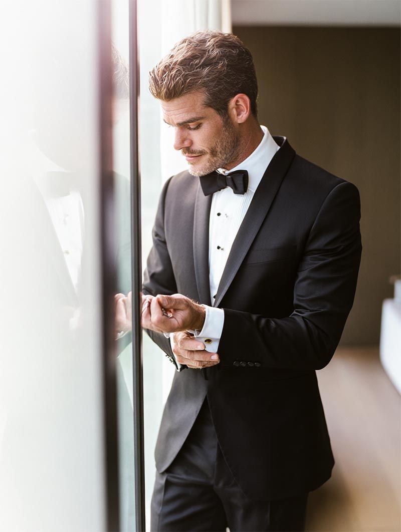 Groom in a tailored black tuxedo adjusting gold cufflinks in soft window light before his luxury wedding ceremony